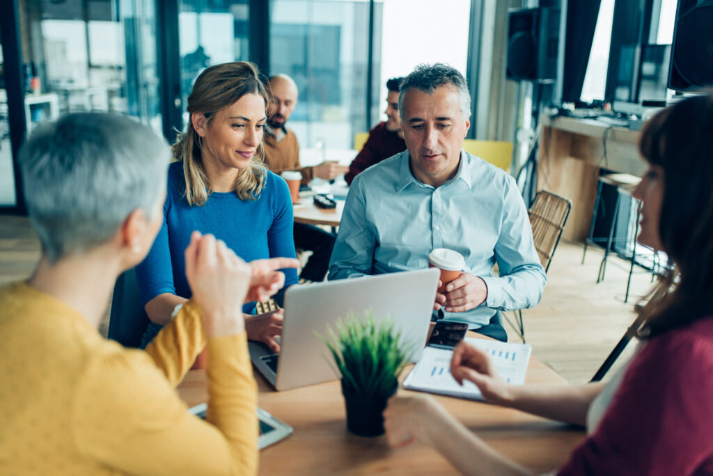 Fyra personer sitter runt ett bord i ett större utrymme. En dator och en krukväxt står på bordet. Personerna samtalar med varandra.
